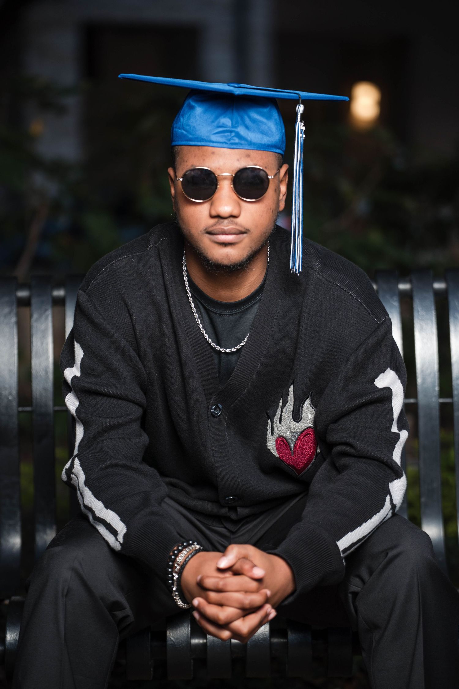 Male graduate in blue cap and sunglasses sits on bench with hands clasped in dark setting.