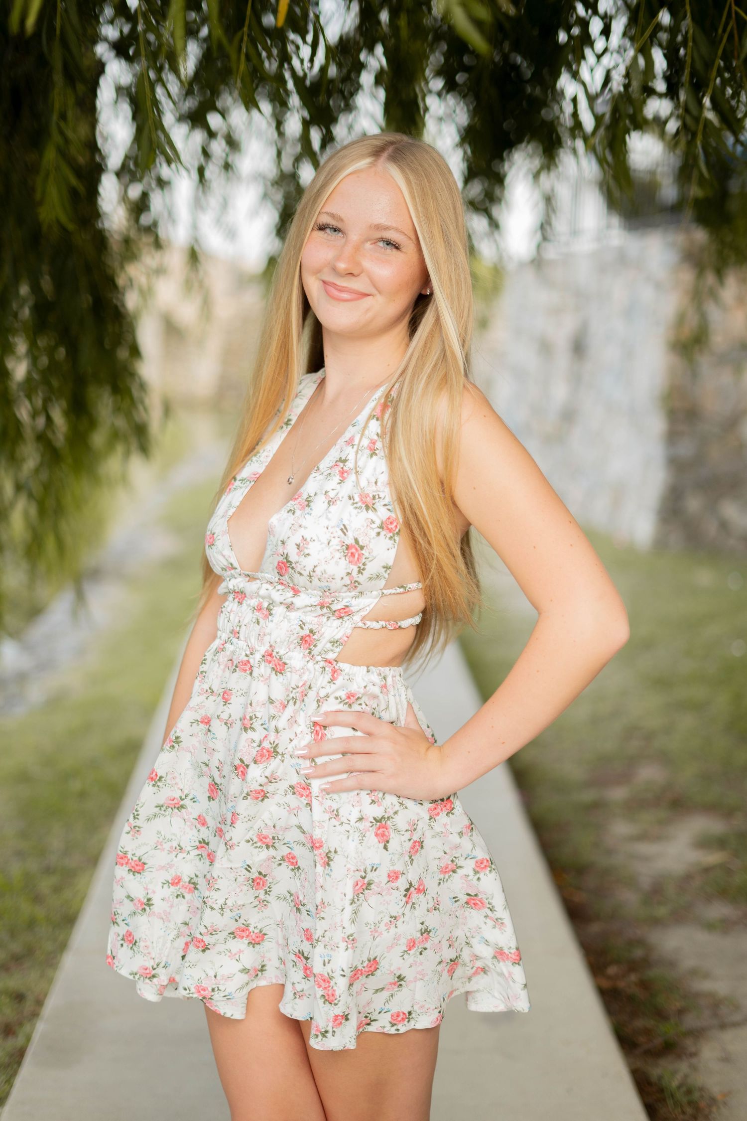 Smiling blonde teenage girl in white floral dress poses outdoors under weeping willow branches.