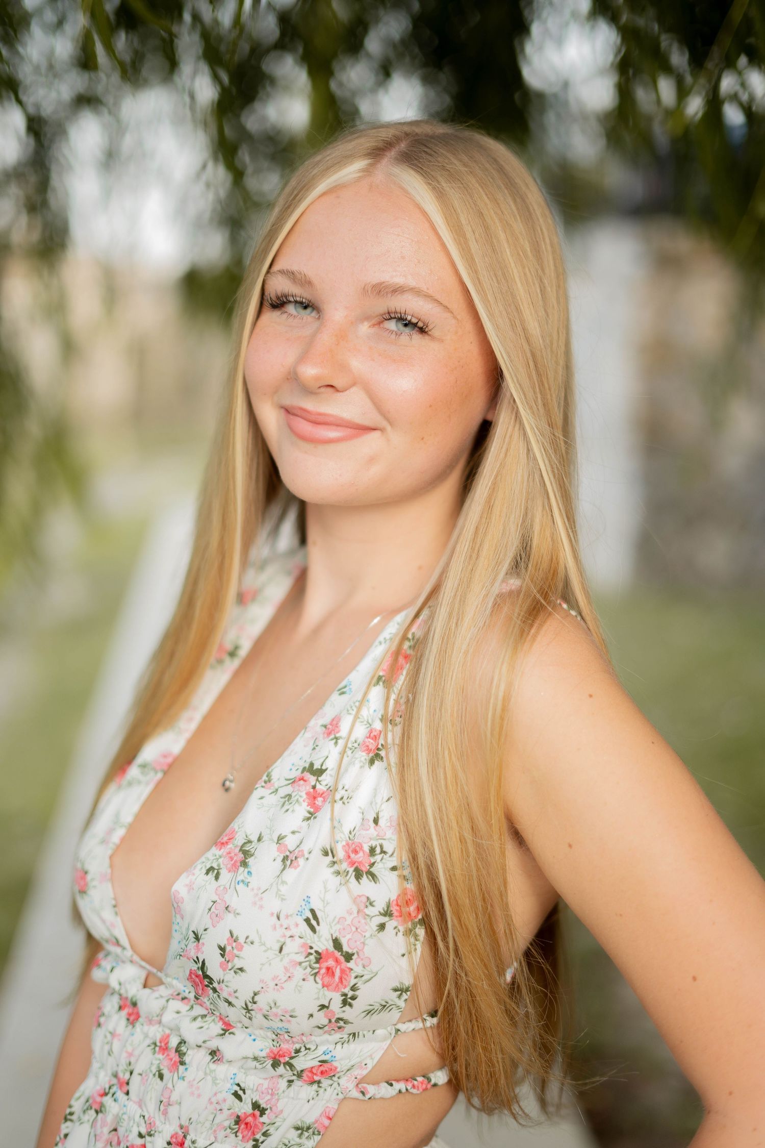 Smiling young blonde woman in floral dress poses outdoors for senior portrait photo.