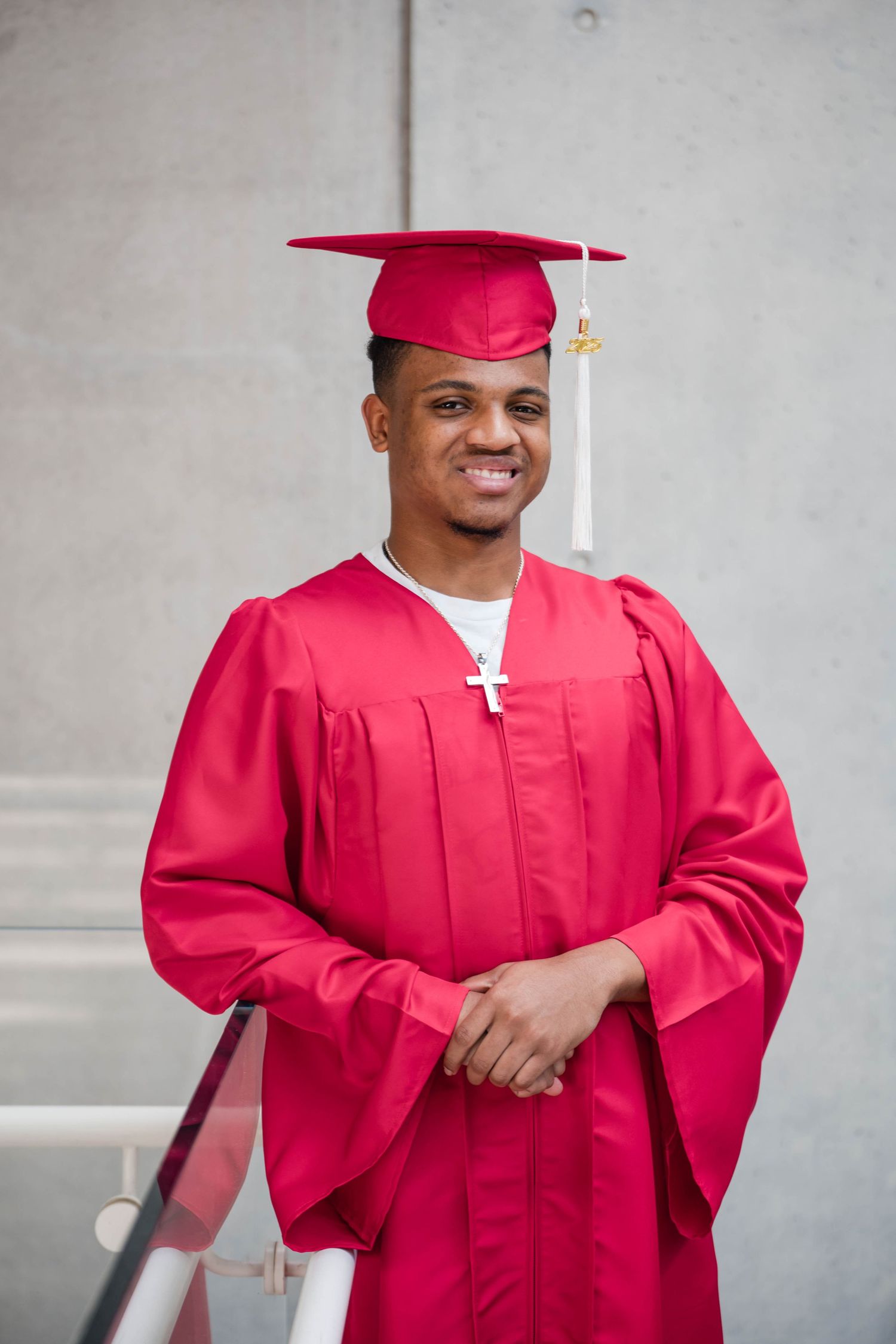 Young man smiling in red graduation cap and gown with cross necklace outdoors.