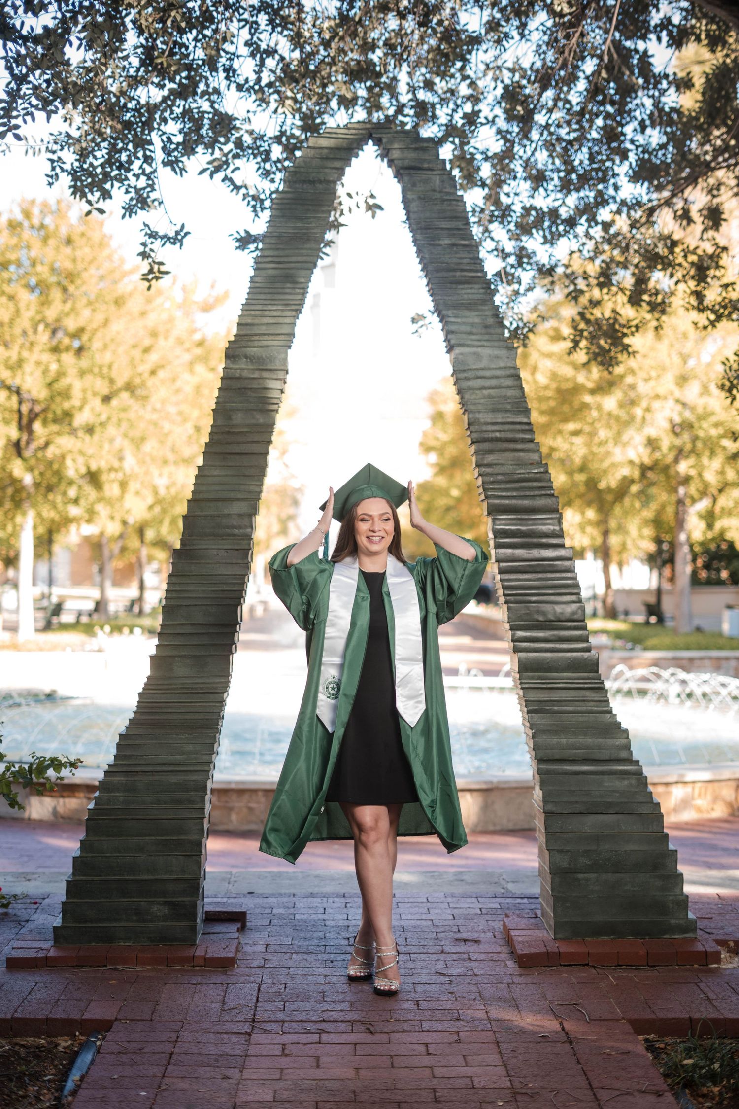 Joyful female graduate in green cap and gown posing under arch sculpture on campus.