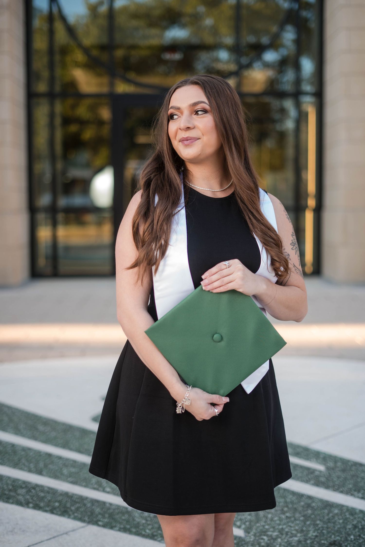 Young woman in black dress holding green graduation cap outside modern building.