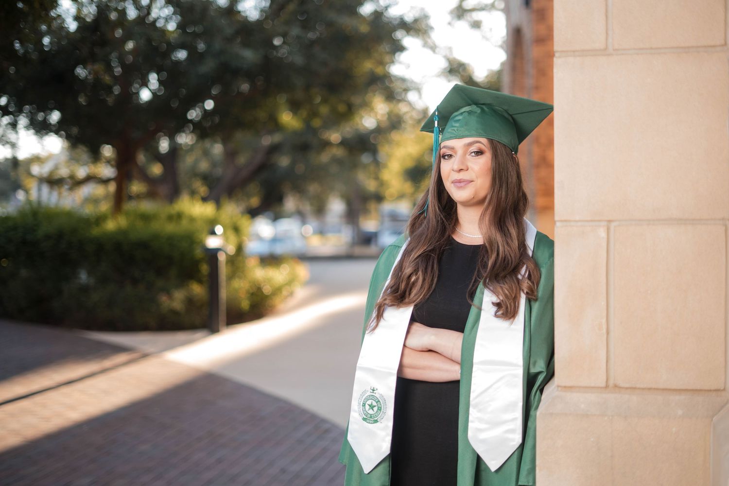 Female graduate in green cap and gown leaning against column on college campus.