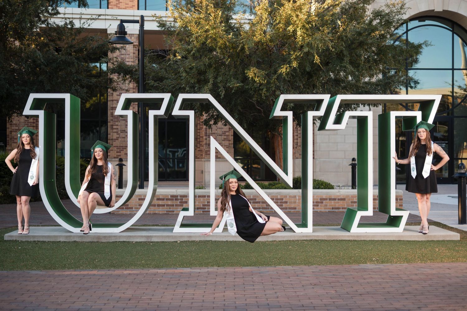 Four female graduates pose with large UNT letter sculptures on campus wearing caps and white stoles.