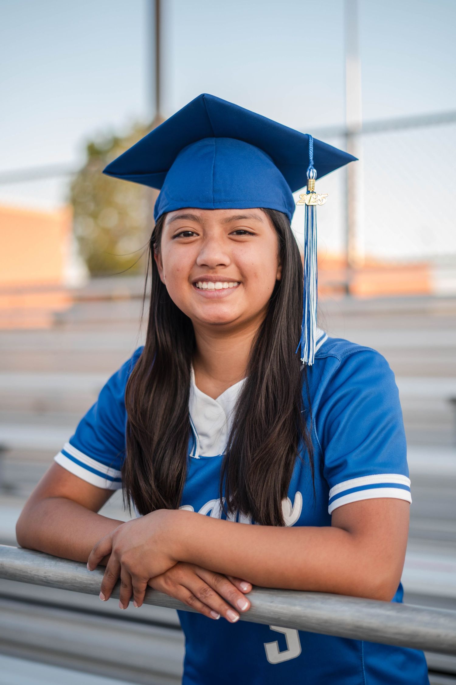 Smiling female graduate in blue cap and Bears jersey leaning on bleachers at sunset.