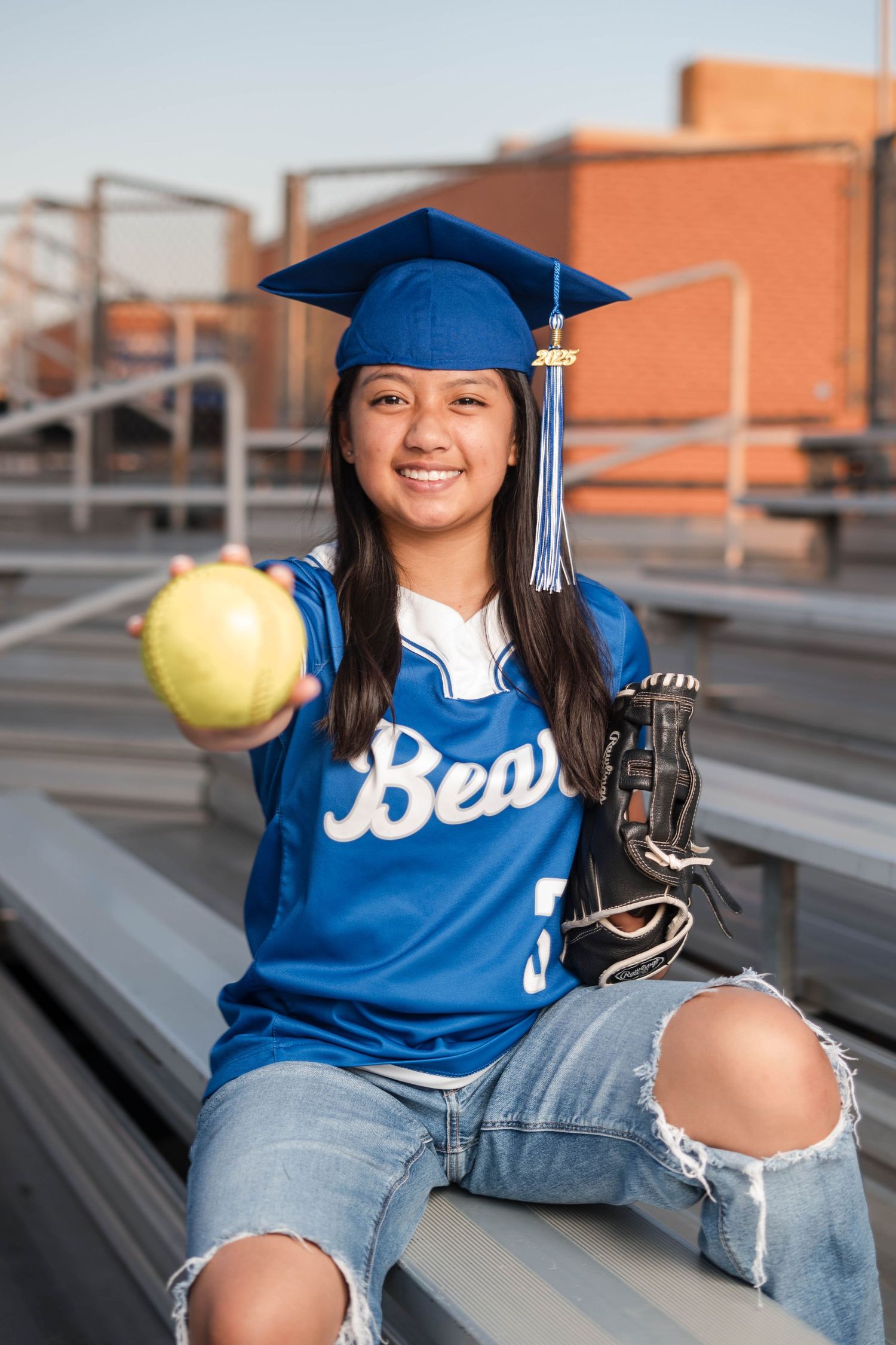 Female graduate in blue cap holding softball while sitting on bleachers in Bears jersey.
