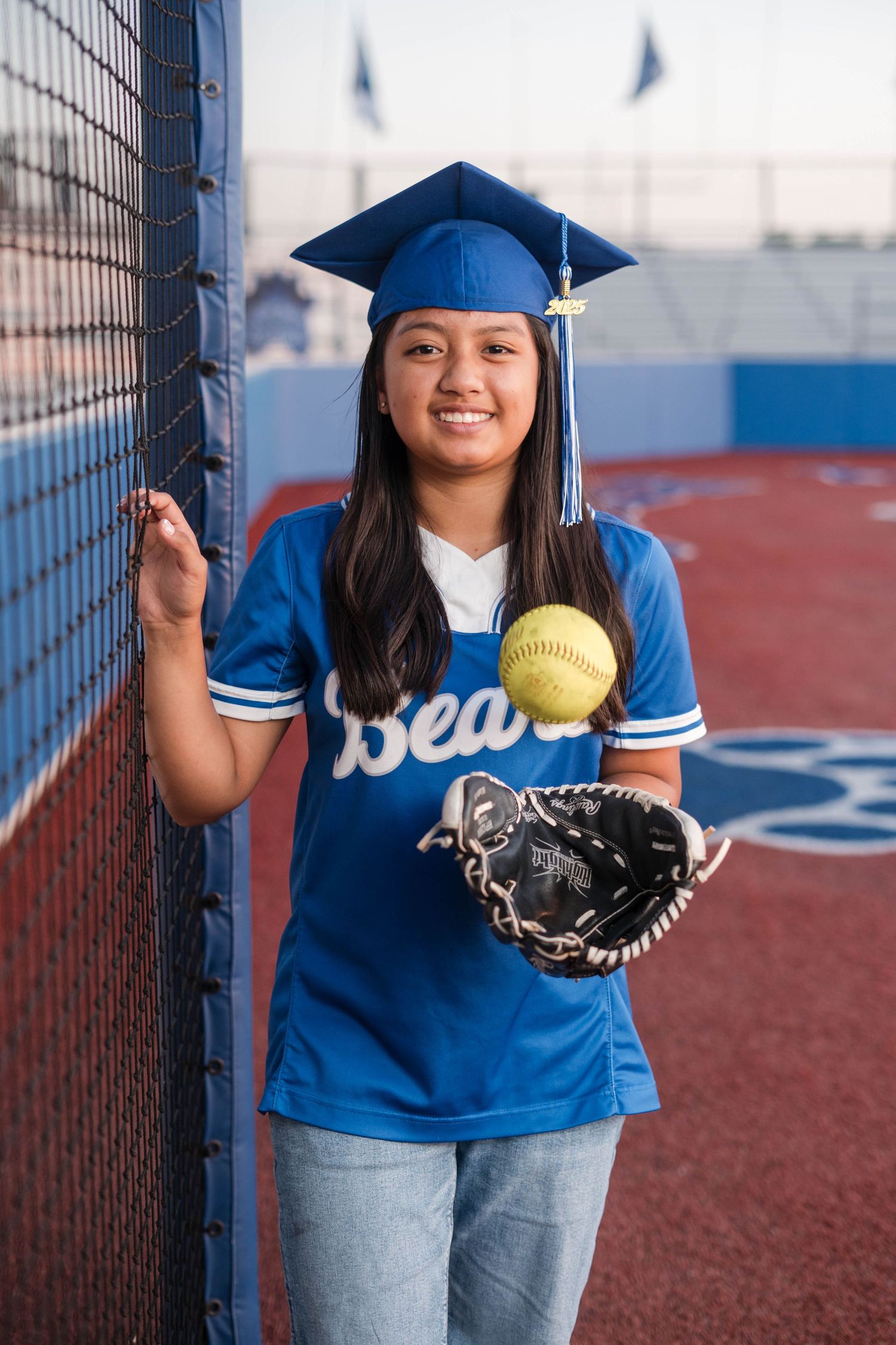 Female graduate in blue cap and Bears jersey holding softball glove by field fence.