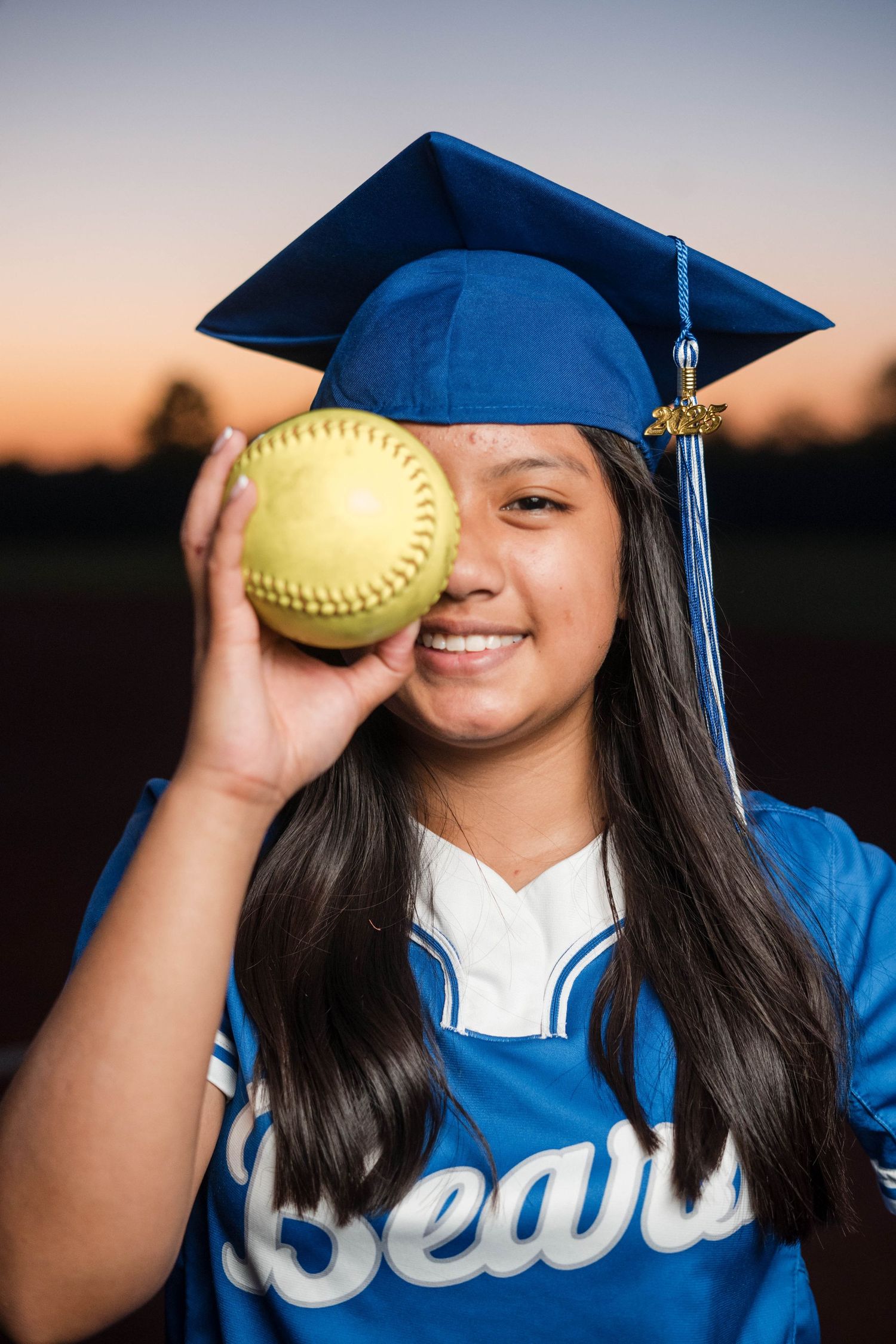 Female graduate in blue Bears cap playfully covering eye with yellow softball at dusk.