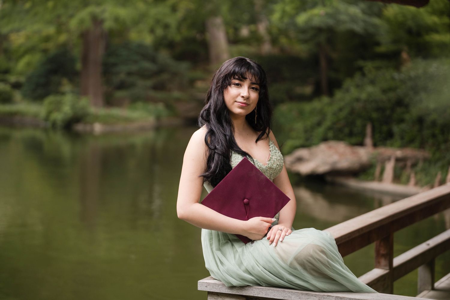 Young woman in green dress holding burgundy diploma folder by a serene garden pond.