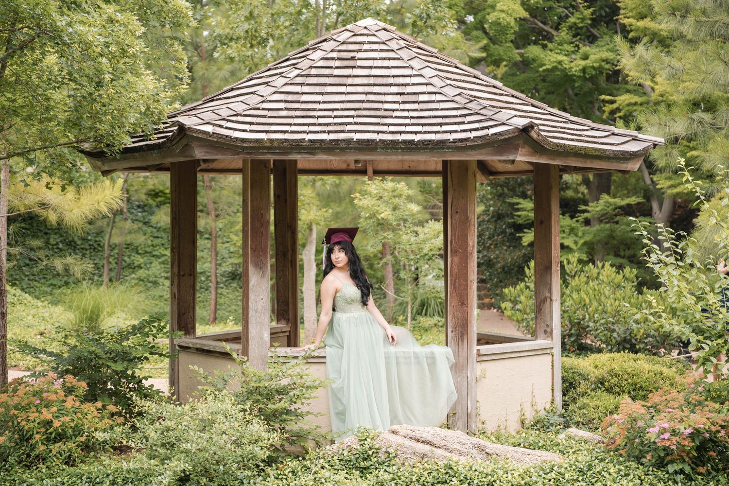 Graduate in mint green dress standing in rustic wooden gazebo surrounded by greenery.