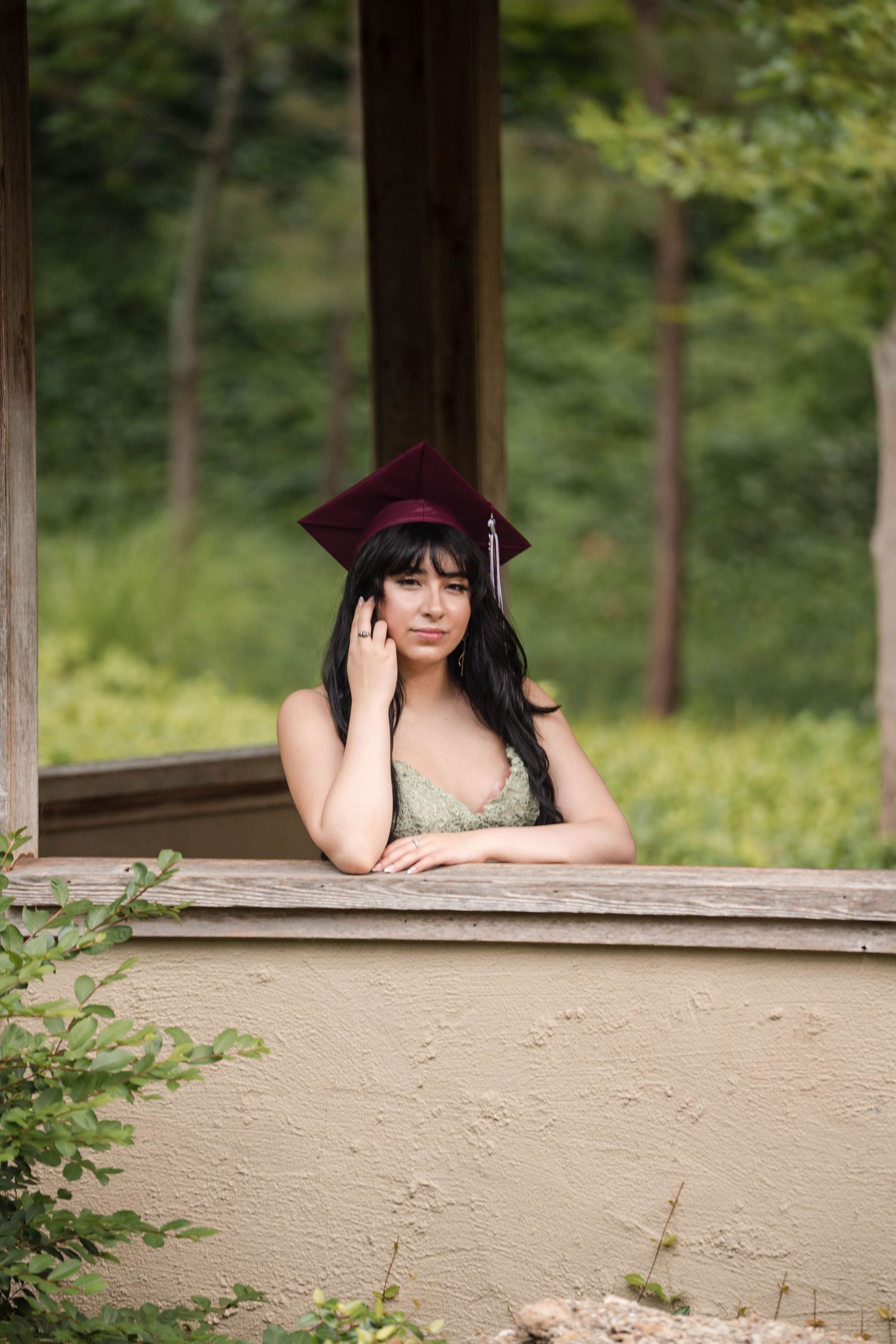 Female graduate in burgundy cap leaning on gazebo railing in a garden.