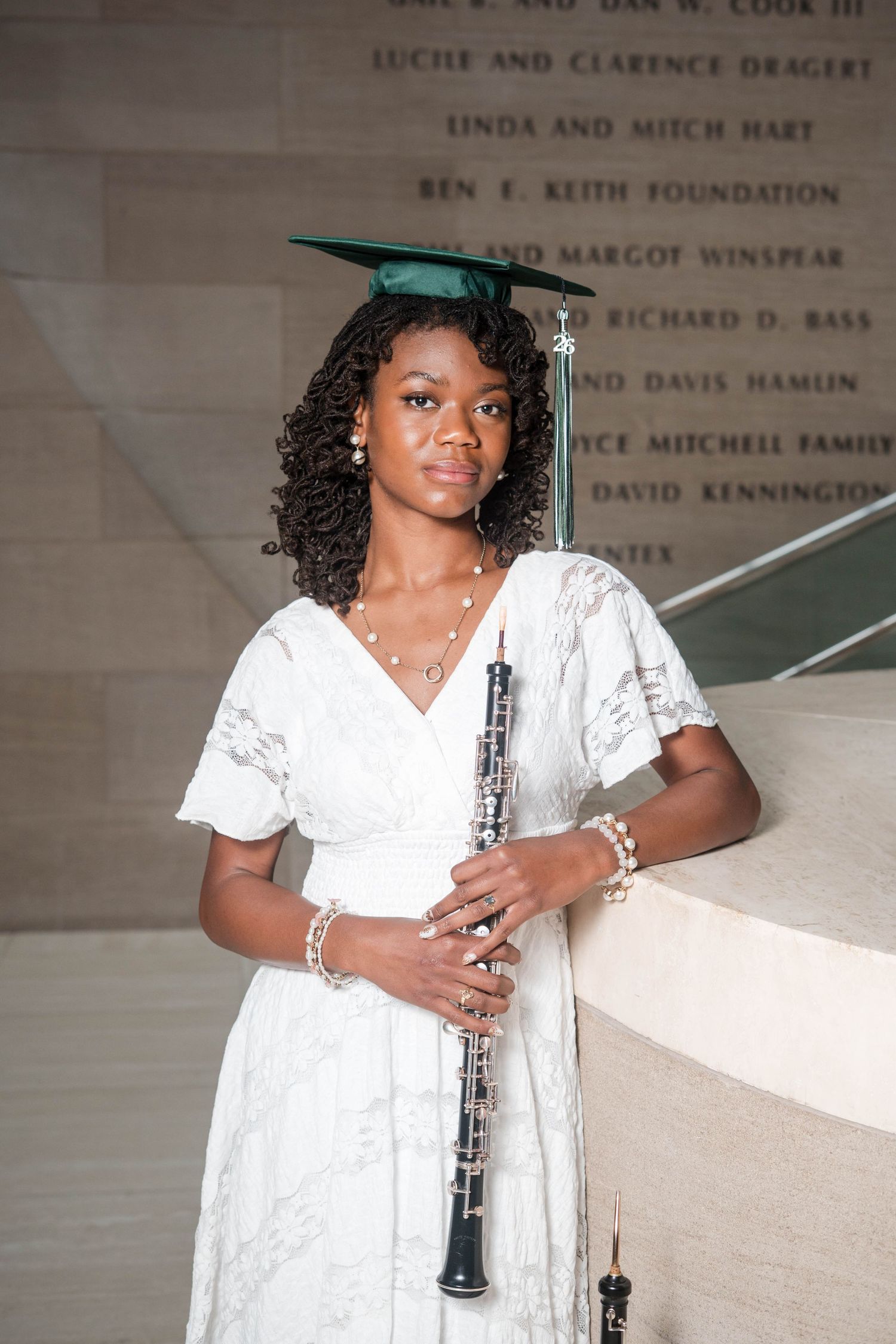 Young woman in white dress and green graduation cap holds a clarinet, posing proudly for senior graduation photos.