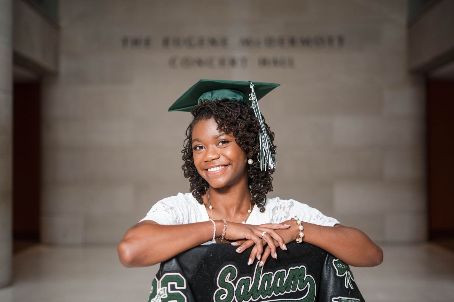 Smiling graduate in green cap poses with Salaam jacket at Eugene McDermott Concert Hall.