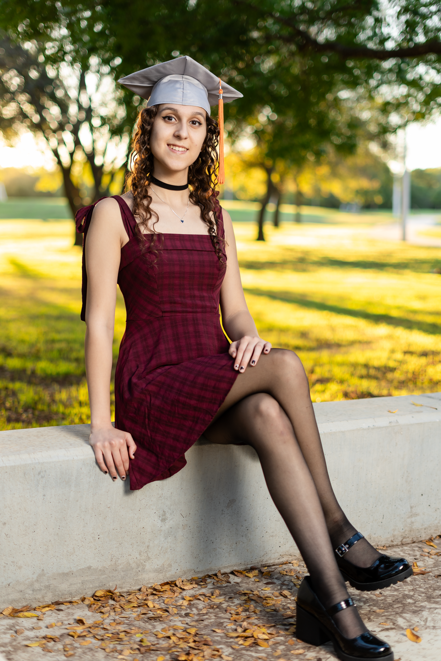Graduate in burgundy dress and cap sitting on concrete ledge in a sunlit park setting.