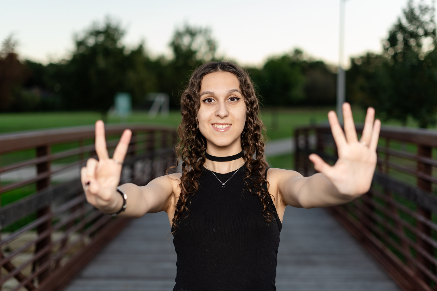 Curly-haired woman in black top showing seven fingers while standing on an outdoor bridge.