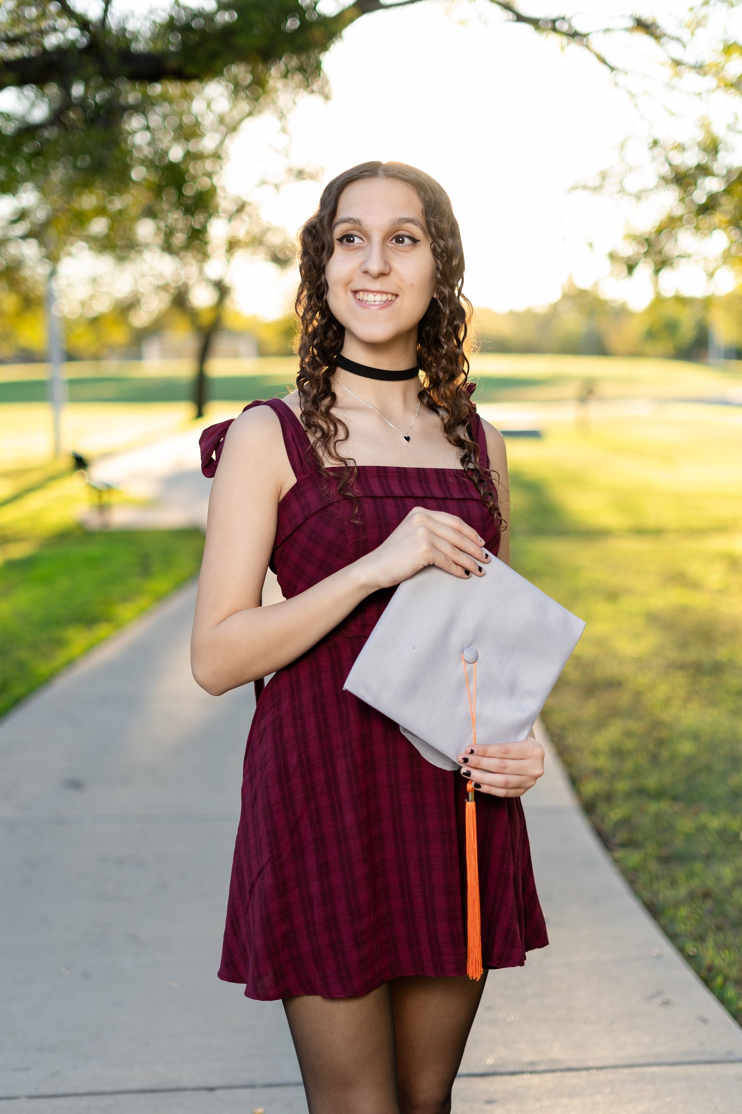 Young woman in burgundy dress holding gray graduation cap on a sunny outdoor walkway.