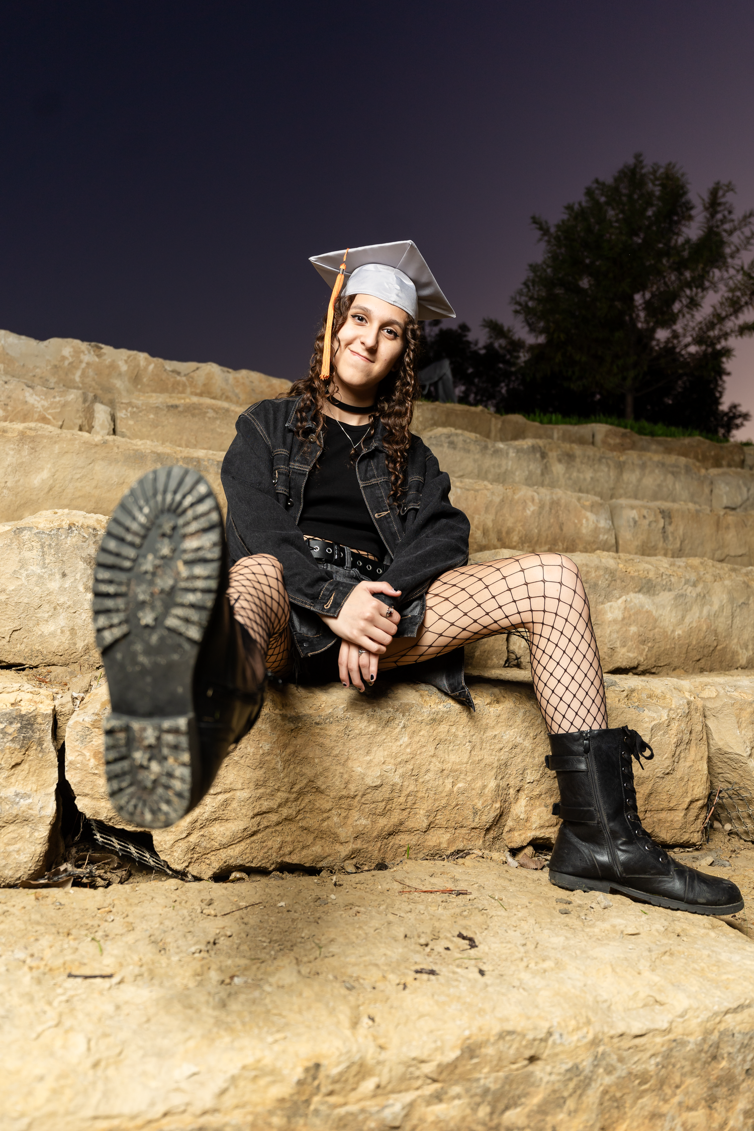 Graduate in cap, denim jacket, fishnet tights, and boots sitting on stone steps at night.