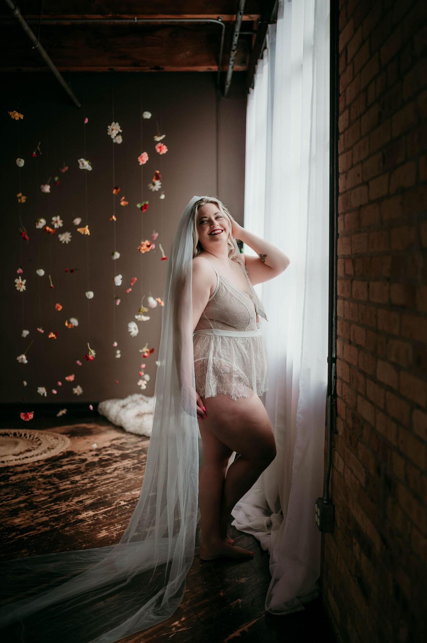 Romantic boudoir photoshoot with flowing white fabric and falling rose petals in moody lighting against brick wall.