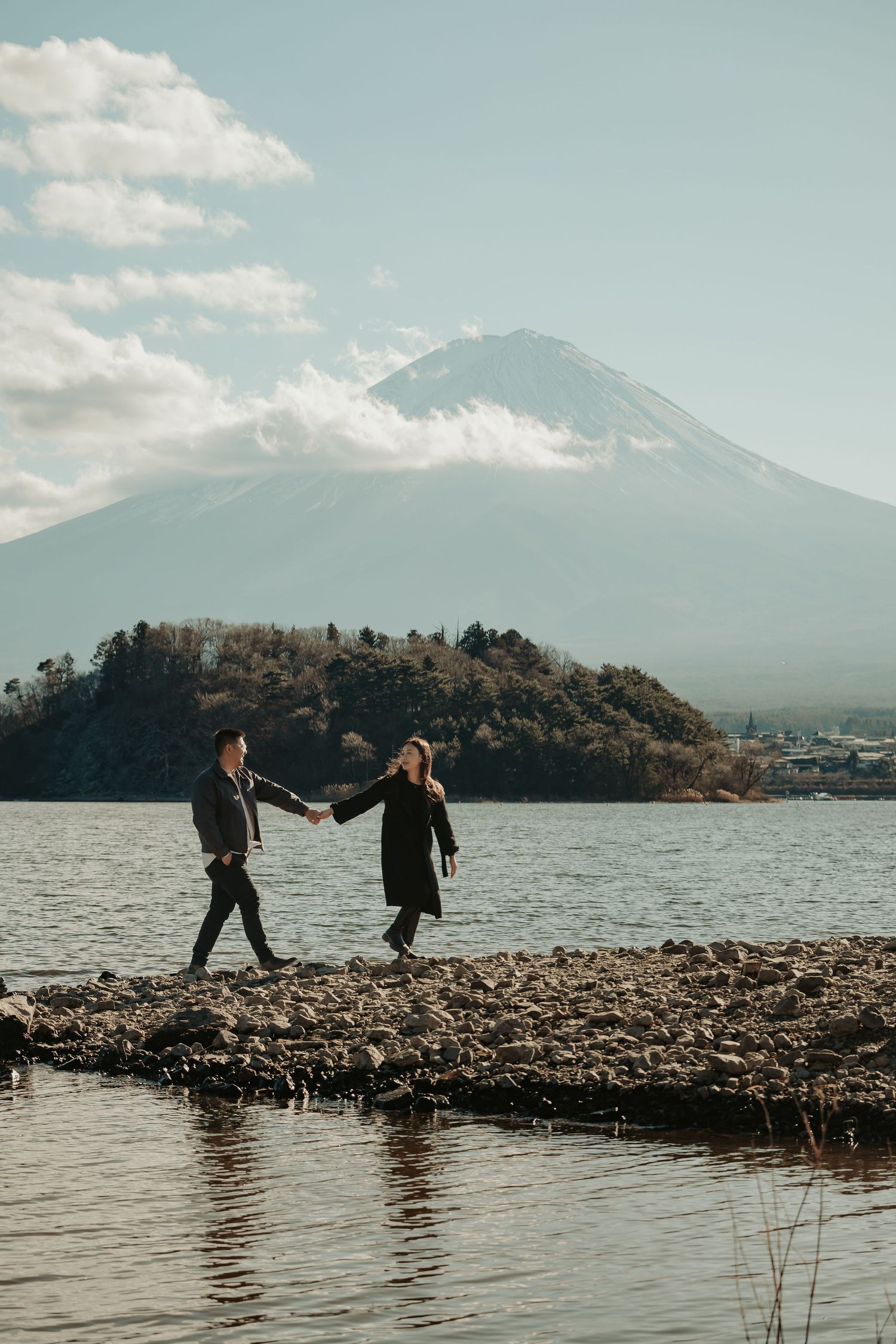 Two people walk hand in hand along a rocky lakeshore with majestic Mount Fuji rising in the misty background.