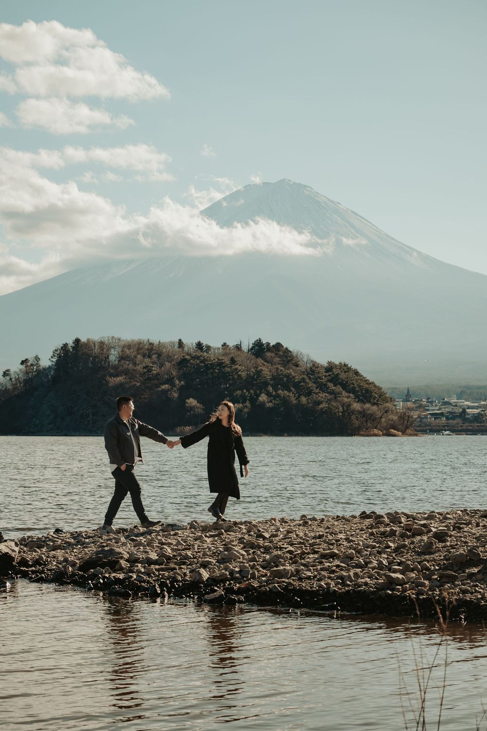 Two people walk hand in hand along a rocky lakeshore with majestic Mount Fuji rising in the misty background.