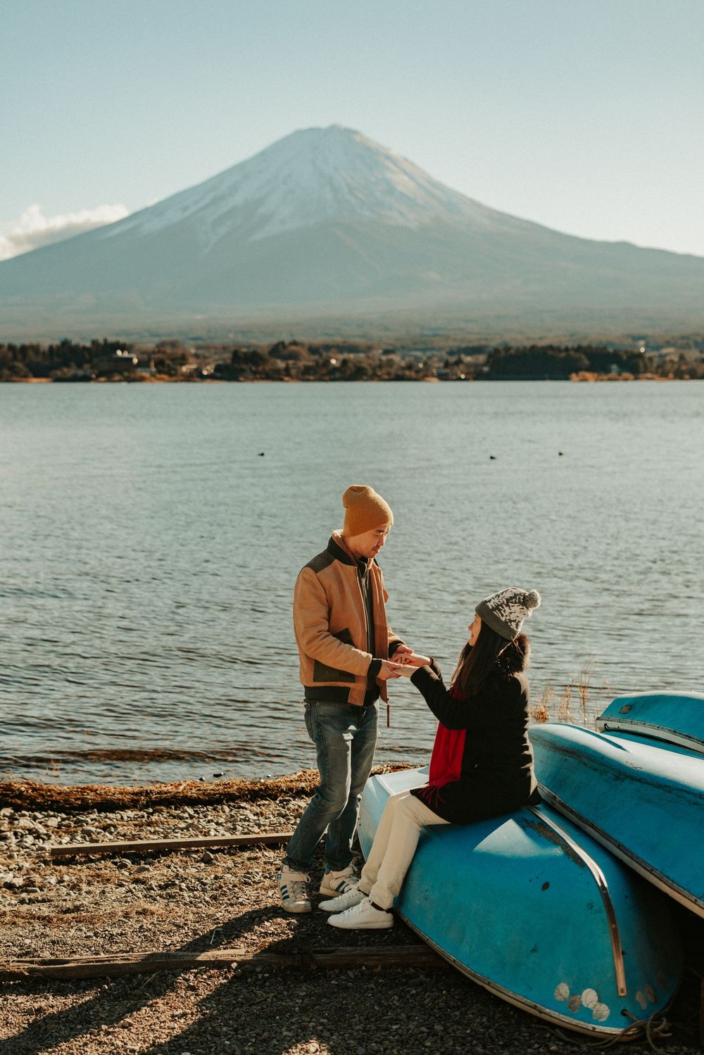Two people sit by a tranquil lake with Mount Fuji visible in the background on a calm evening.