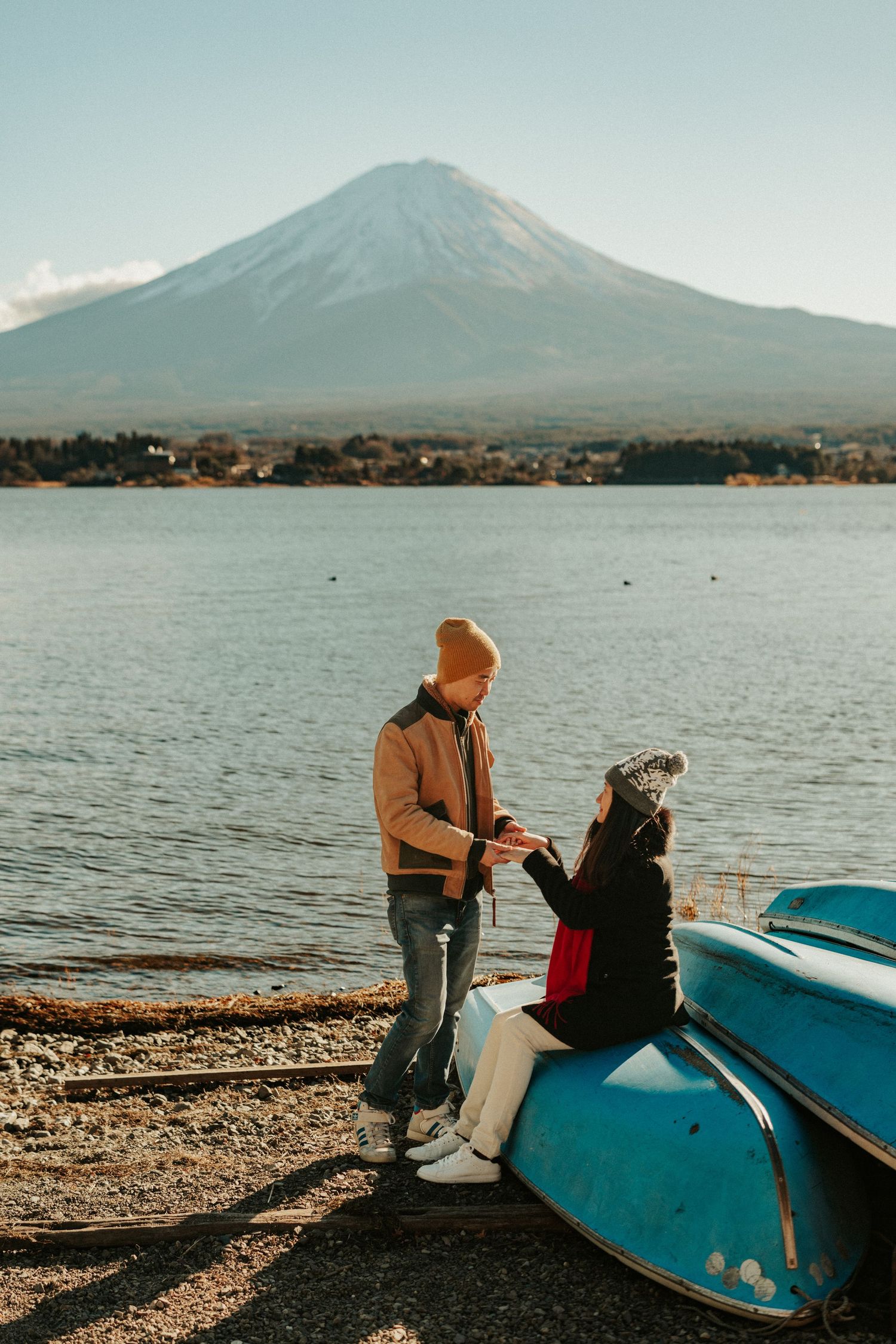 Two people sit by a tranquil lake with Mount Fuji visible in the background on a calm evening.