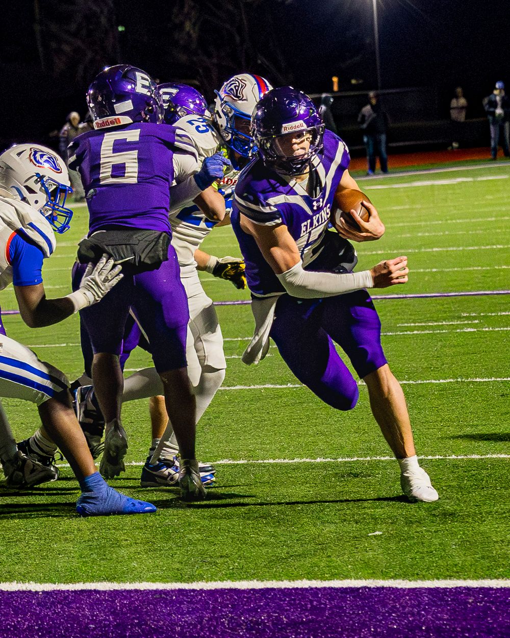 Elkins Varsity Football player in purple and white uniforms scores a touchdown in an intense play during a nighttime game on the field.