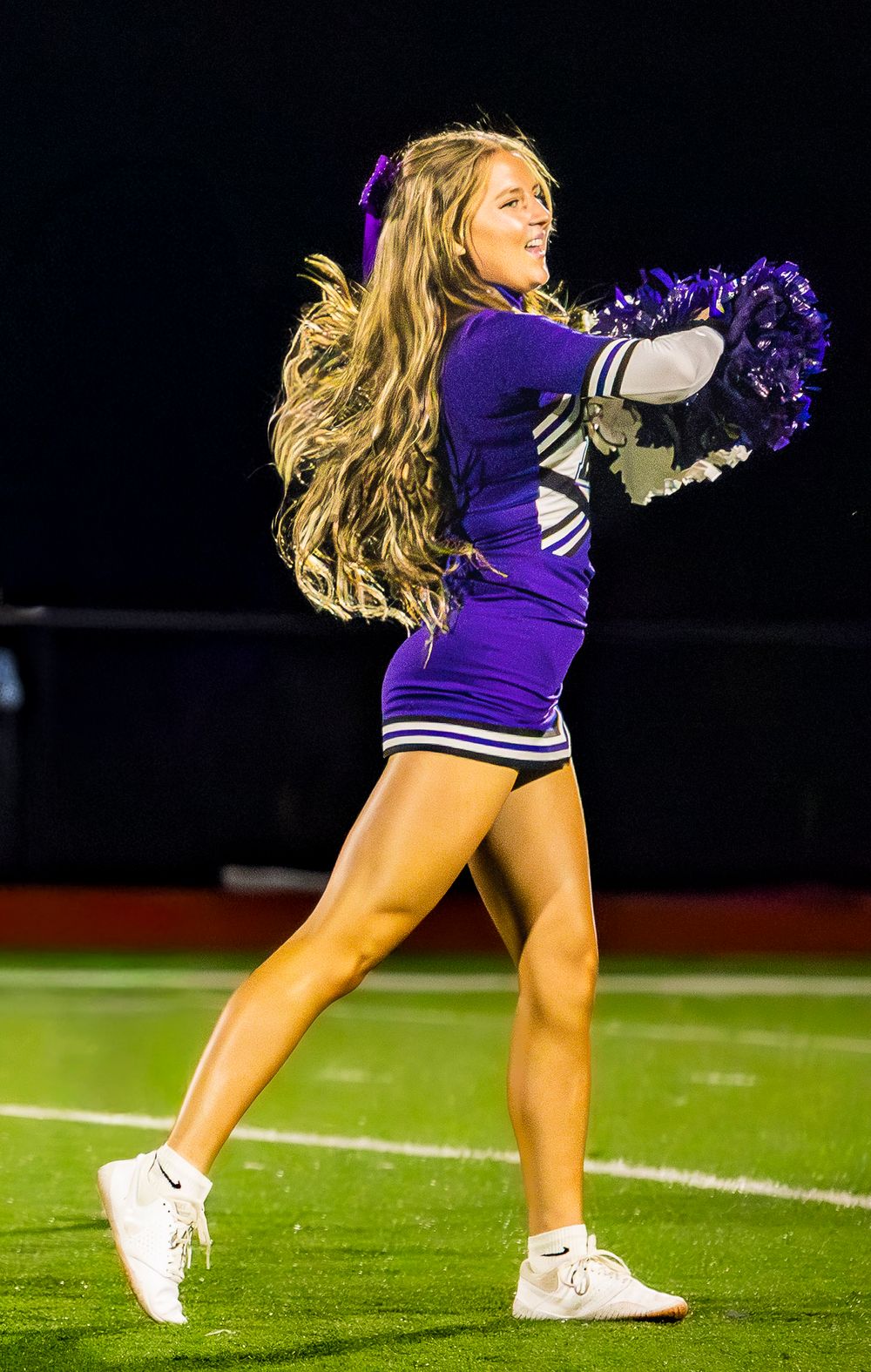 Elkins AR cheerleader performs a dynamic dance routine in a purple uniform on a football field at night.