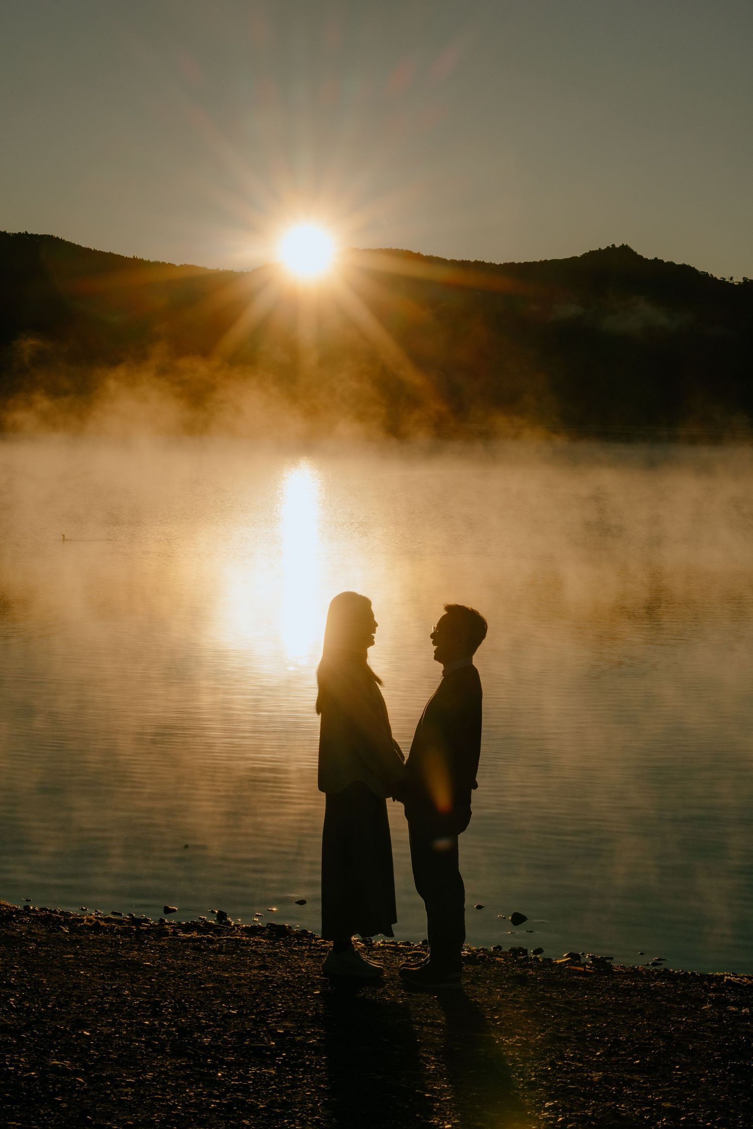 Silhouettes of figures stand by a misty lake at sunrise with sunlight reflecting dramatically on the water's surface.