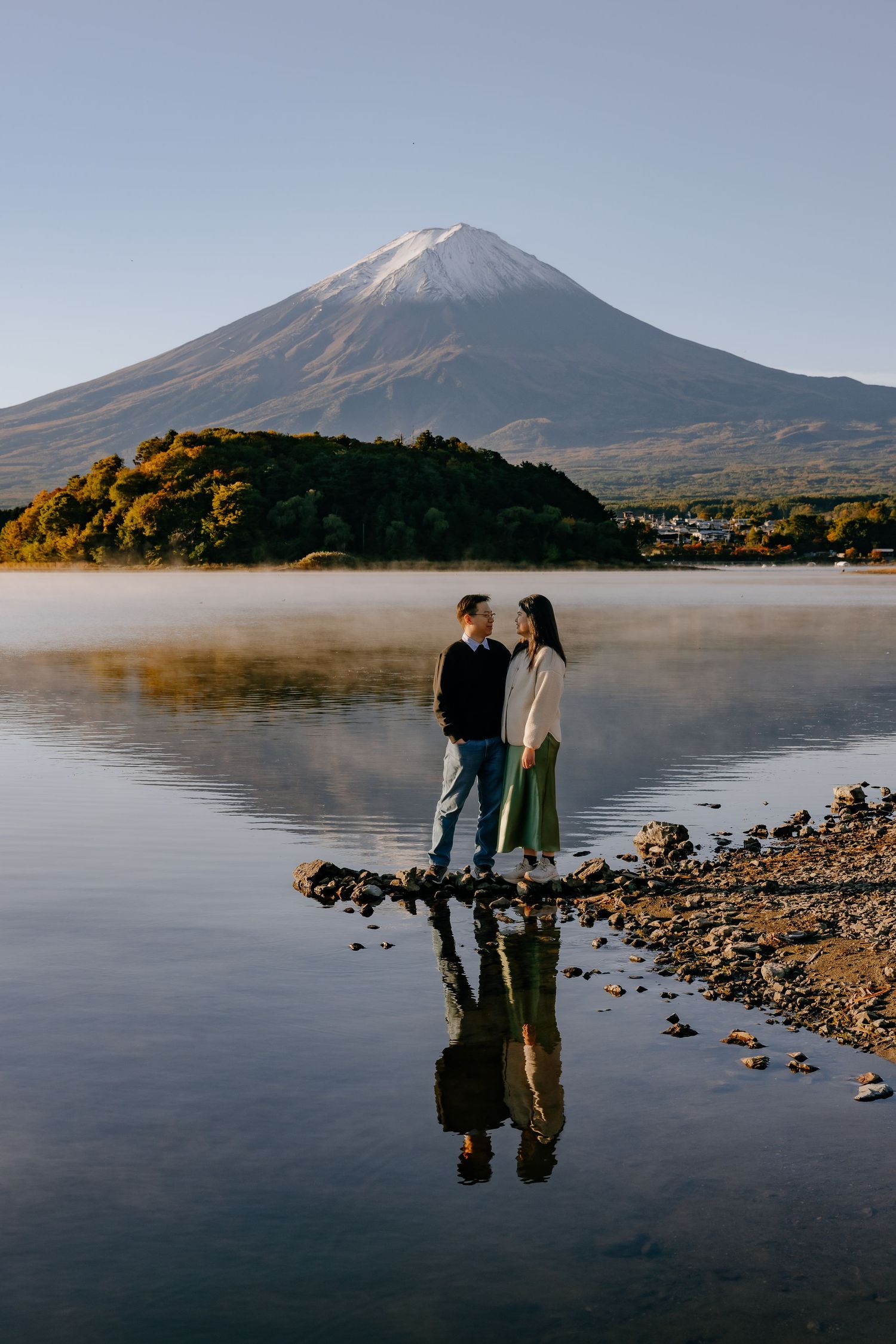 Two people stand on a rocky lakeshore with Mount Fuji rising majestically in the background on a misty morning.