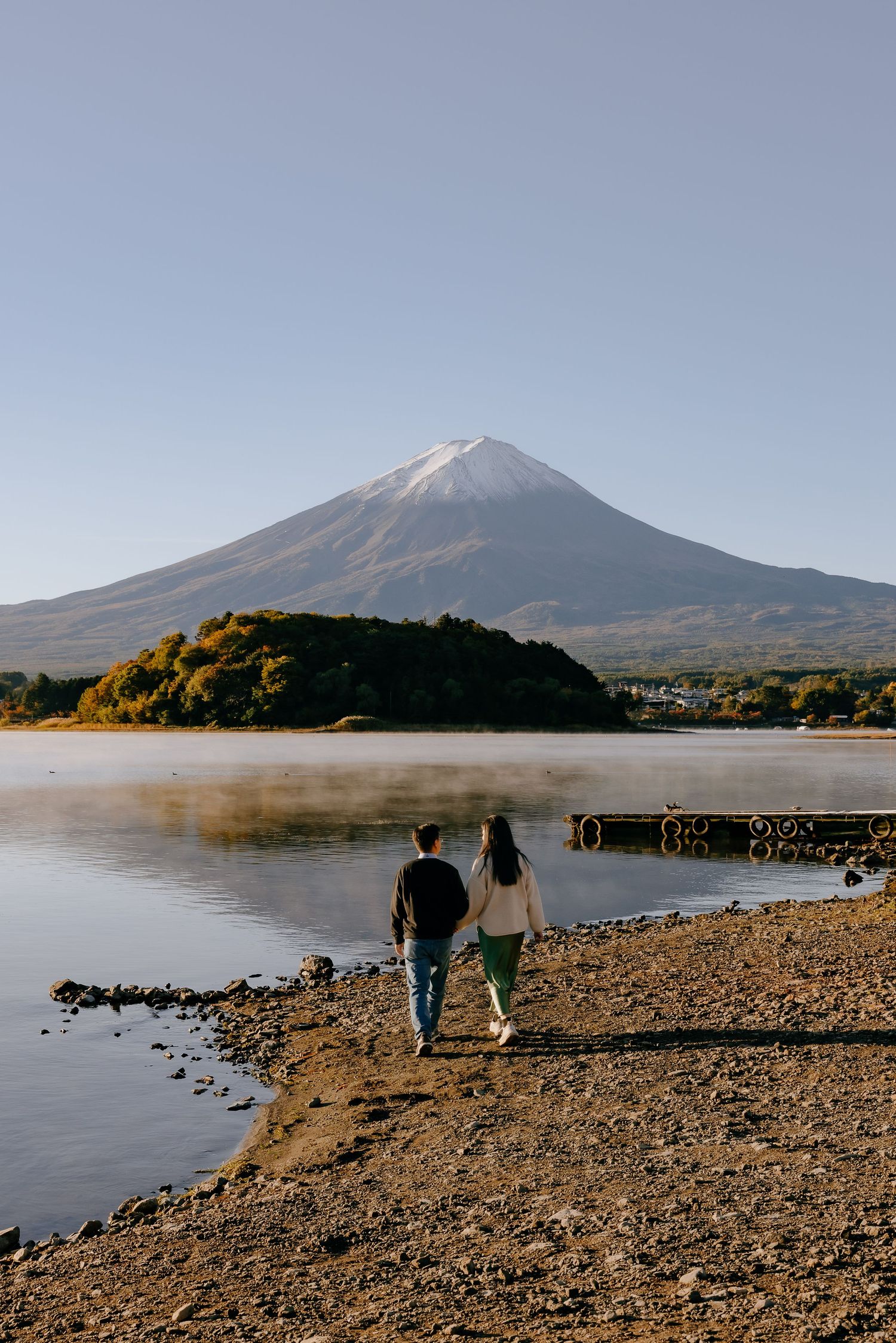 Two people walk along the rocky shoreline of Lake Yamanaka with Mount Fuji looming in the misty background.