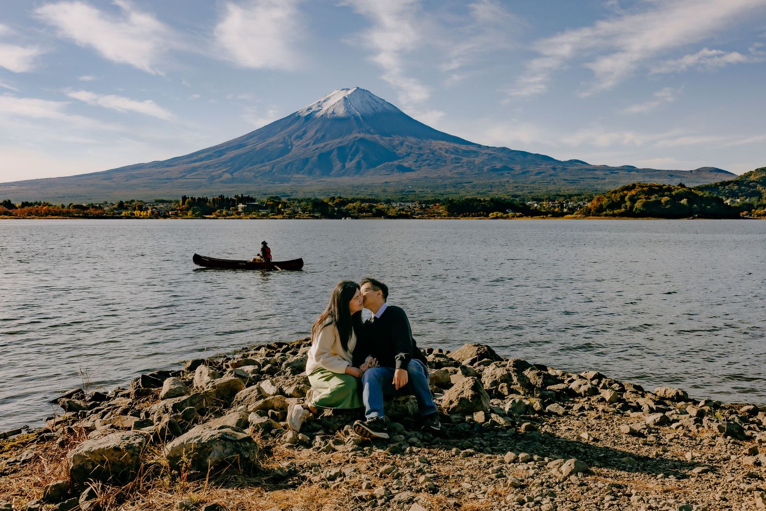 A scenic view of Mount Fuji with a lake in the foreground, featuring kayaking and rocky shoreline activities.
