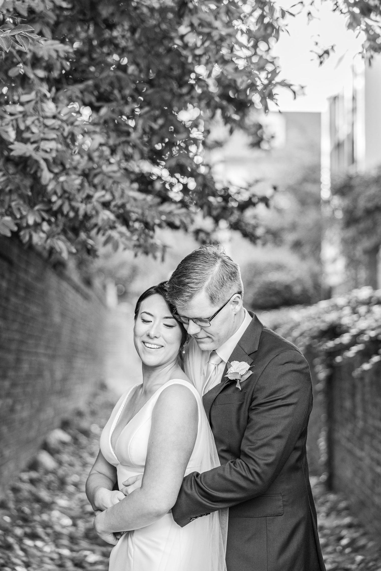 Black and white romantic wedding photo of couple embracing outdoors under tree branches in garden setting.