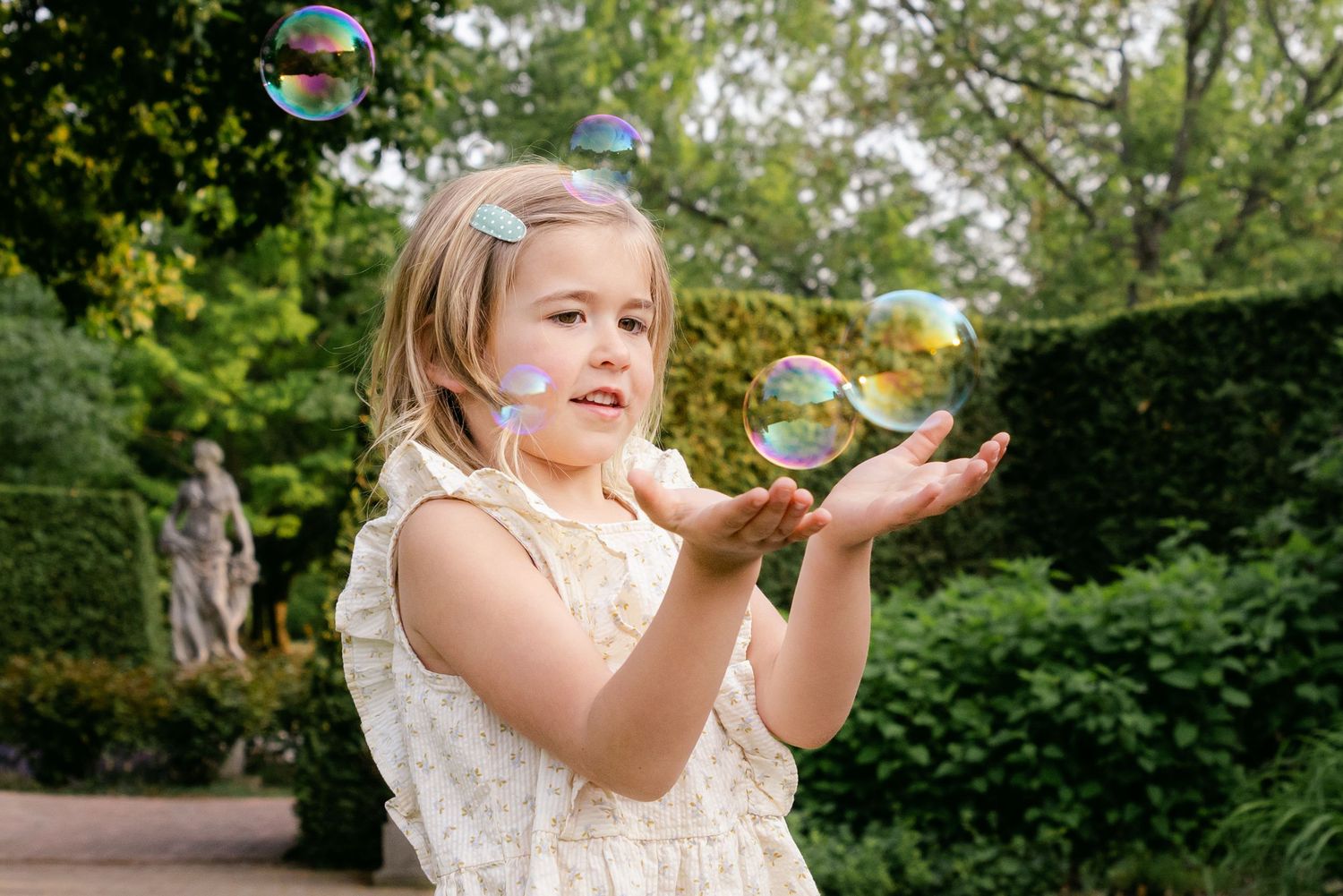 Young girl in floral dress catching soap bubbles in a lush garden with stone statue backdrop.