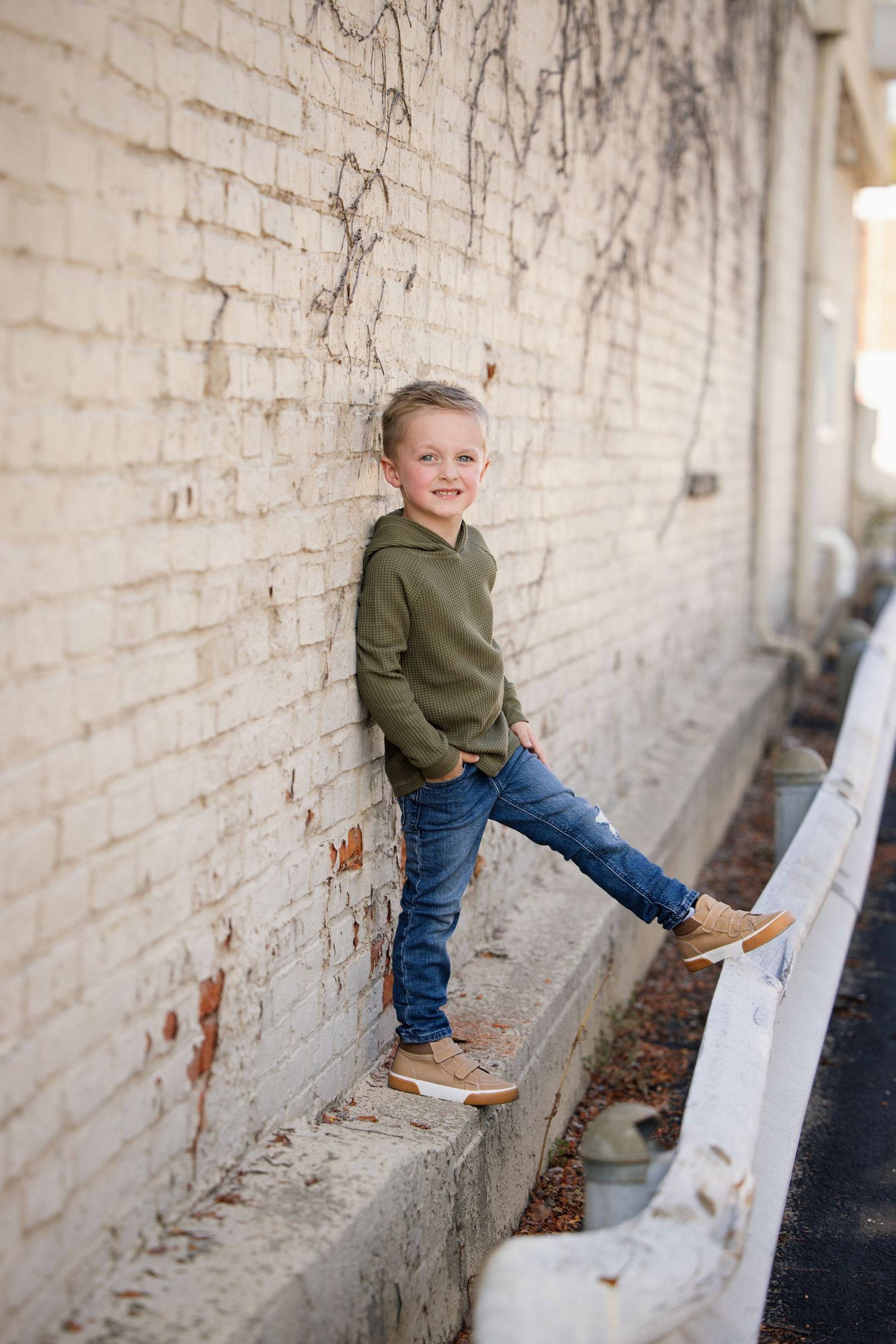 A smiling young boy in a green hoodie and jeans leans against a white brick wall with one foot propped up.
