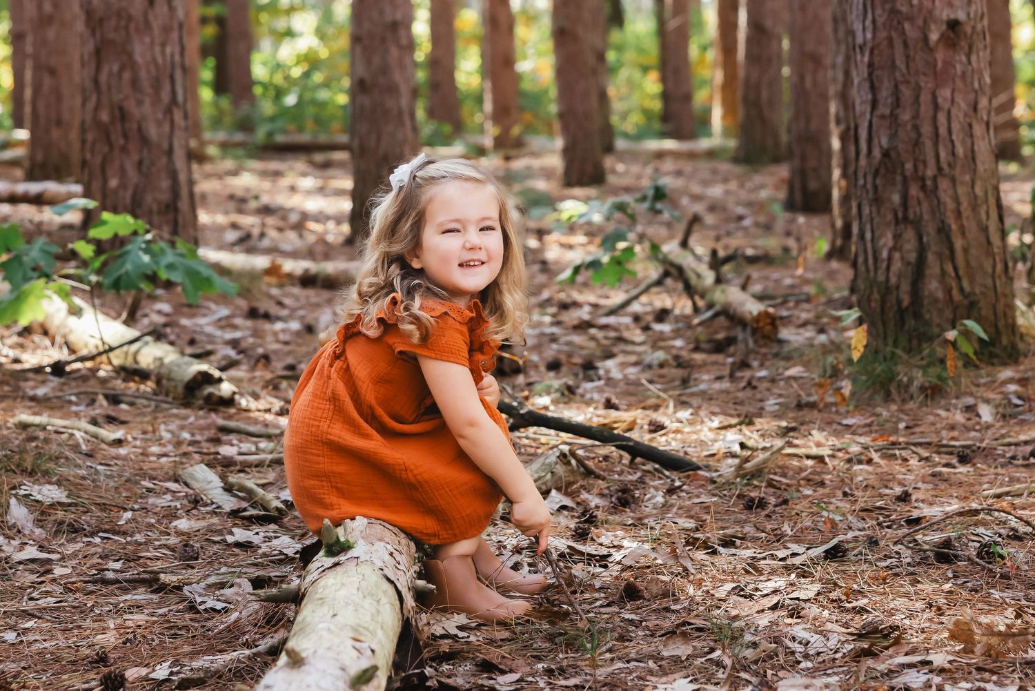 A happy toddler girl in a rust orange dress sits on a log in a sunlit forest, smiling joyfully.