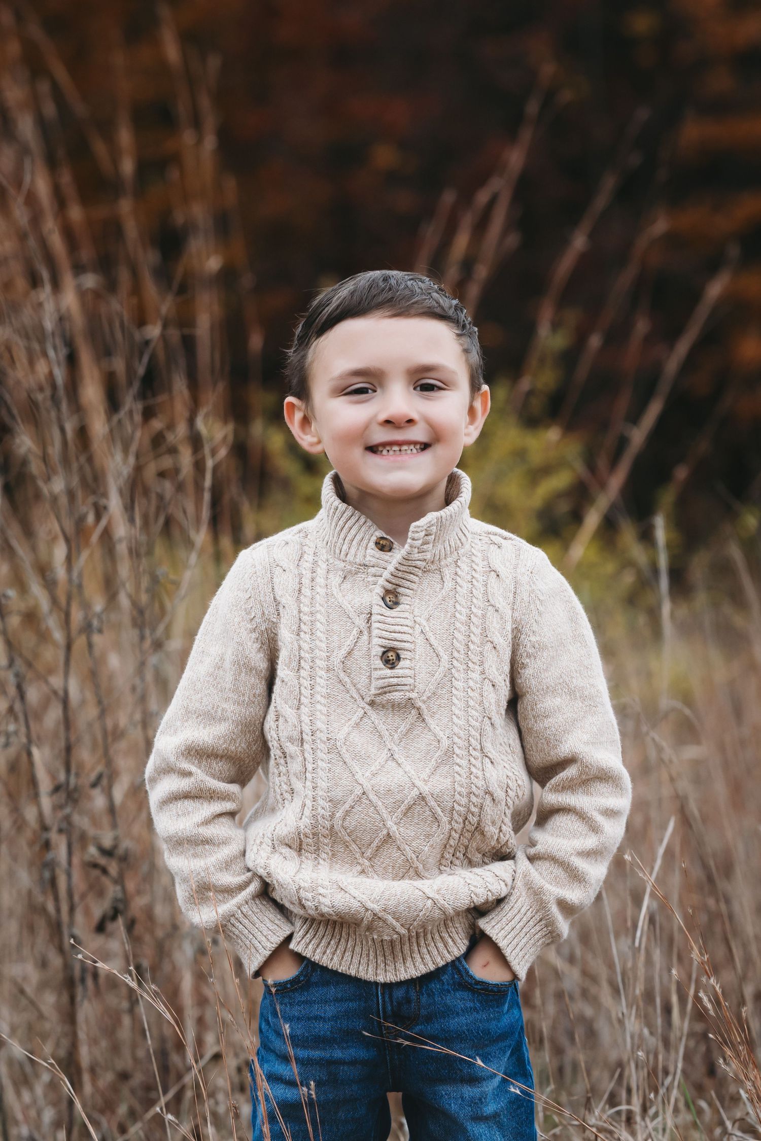 Smiling young boy in a beige cable-knit sweater and jeans standing outdoors in an autumn field.