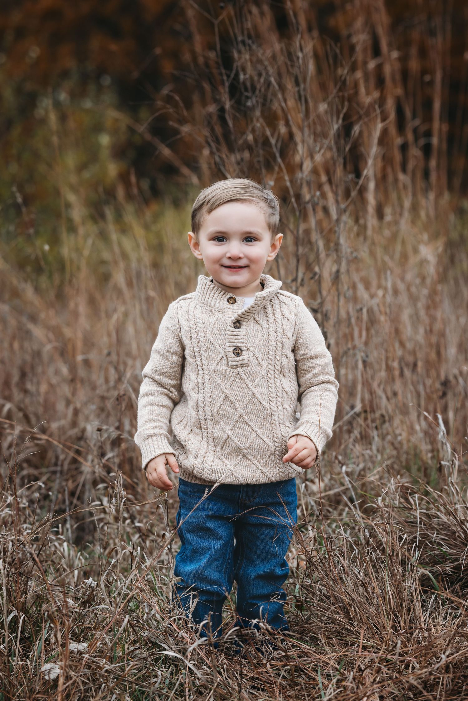 A smiling toddler boy in a cream cable-knit sweater and jeans stands in a field of tall dry grass during autumn.