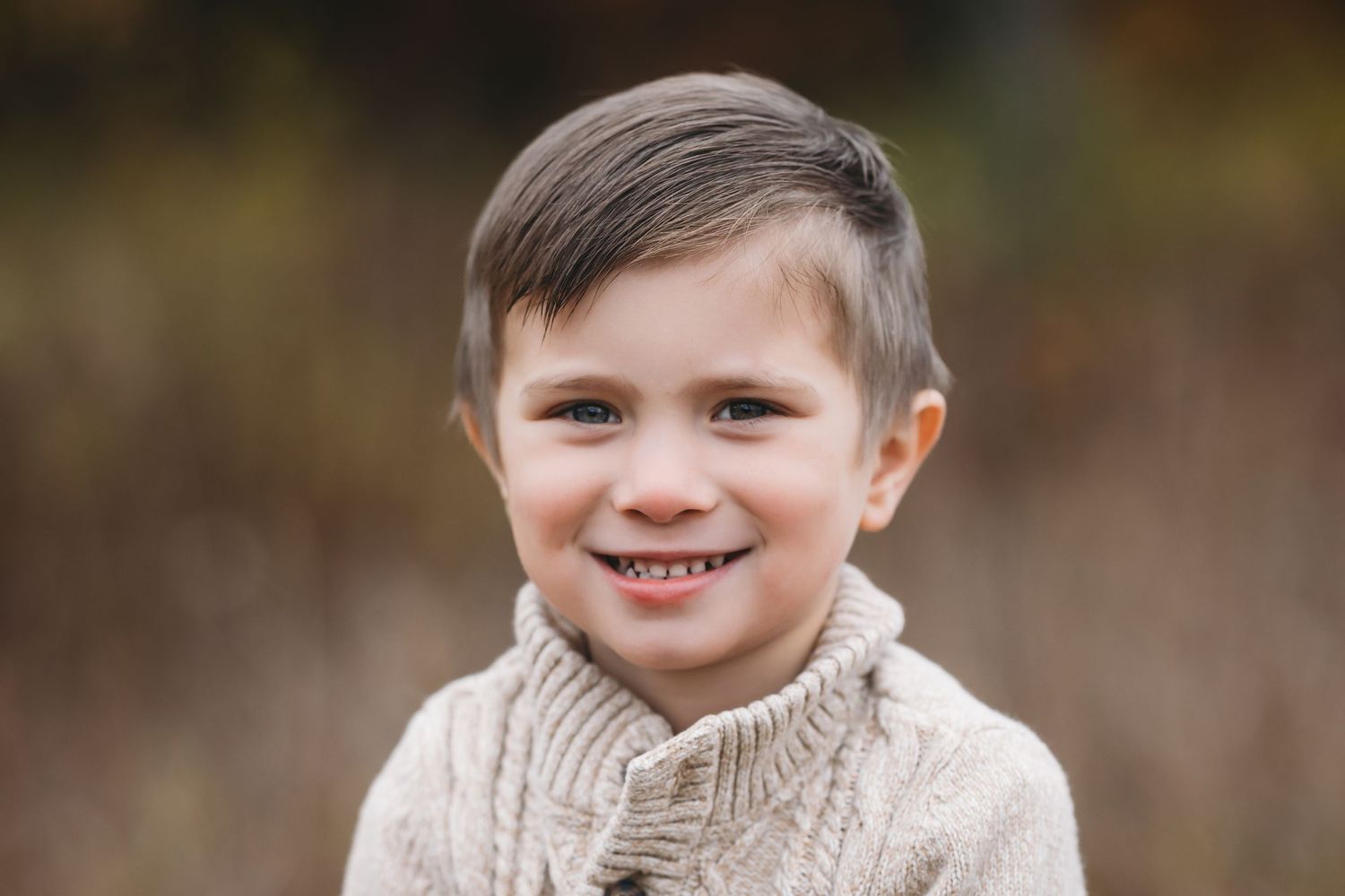 Smiling young boy in a cozy beige cable-knit sweater posing outdoors with a blurred autumn background.