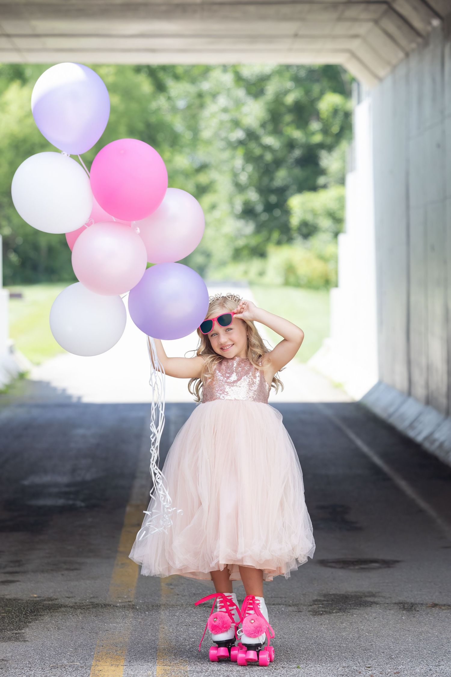 Young girl in pink sequin dress and roller skates holds colorful balloons under a bridge overpass.