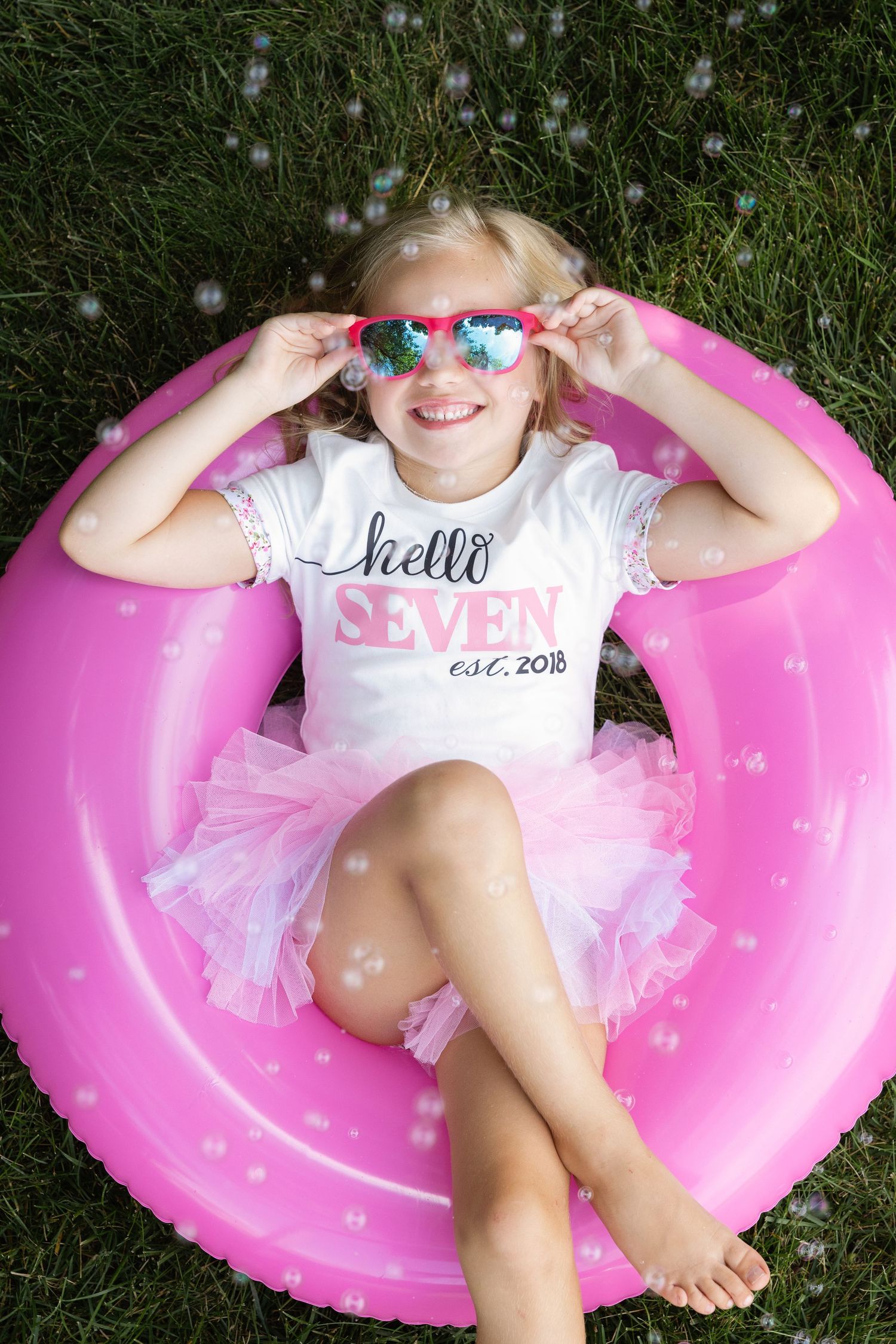 A smiling girl in a Hello Seven birthday shirt and tutu sits in a pink inflatable ring on grass with bubbles around her.