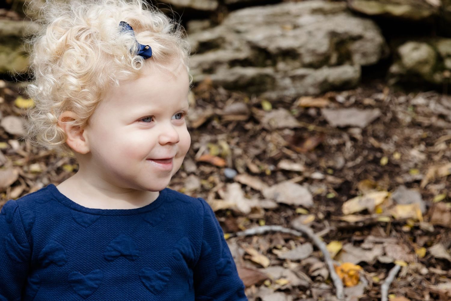 Curly blonde toddler in navy blue shirt smiling outdoors among autumn leaves near stone wall.