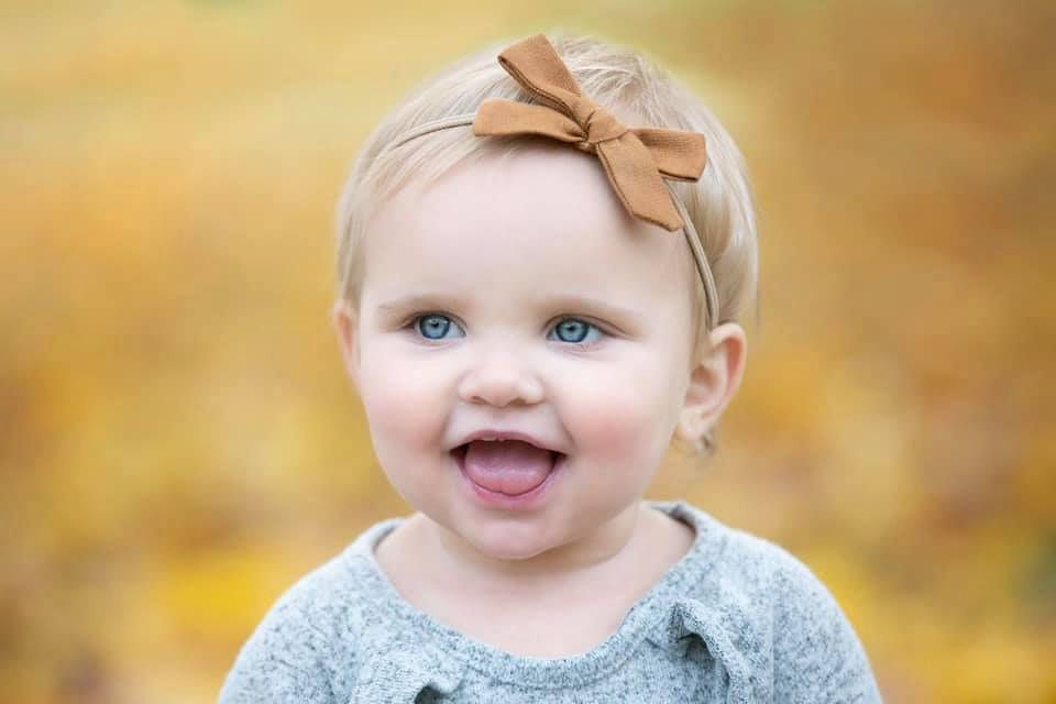 A baby wearing a beige bow headband smiles in a fall setting with golden bokeh background.