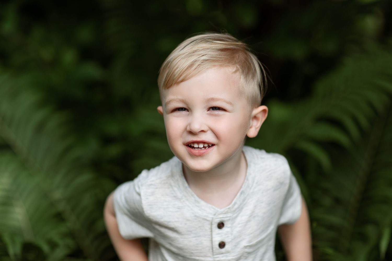 Smiling blond toddler boy in gray shirt sitting outdoors among lush green ferns.