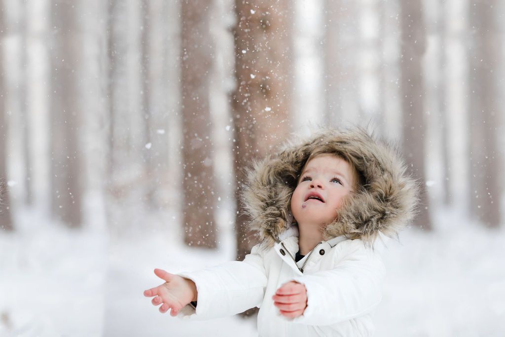 Young child in white fur-hooded jacket catching snowflakes in a snowy winter forest.