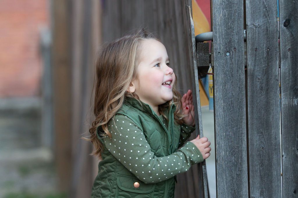 Happy toddler girl in green vest laughing outdoors near wooden fence.