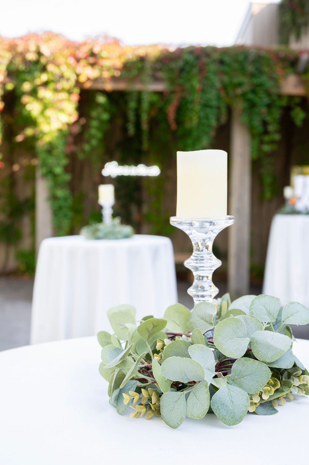 Elegant wedding centerpiece with white candle in crystal holder surrounded by fresh eucalyptus leaves on white tablecloth.