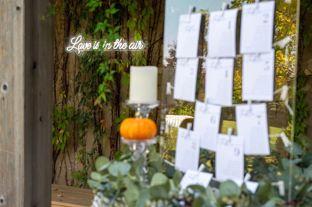A neon 'Love is in the air' sign hangs near a pumpkin and seating chart display at an autumn wedding.