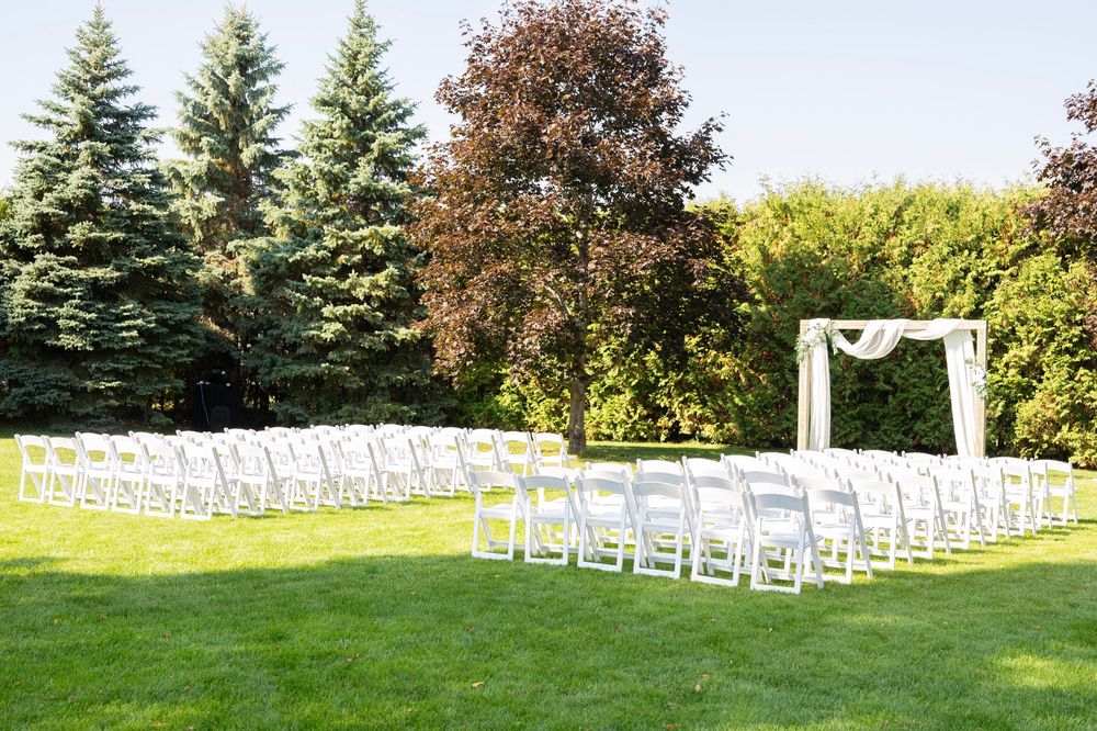 White wedding ceremony chairs arranged in rows on grass with evergreen trees in background.