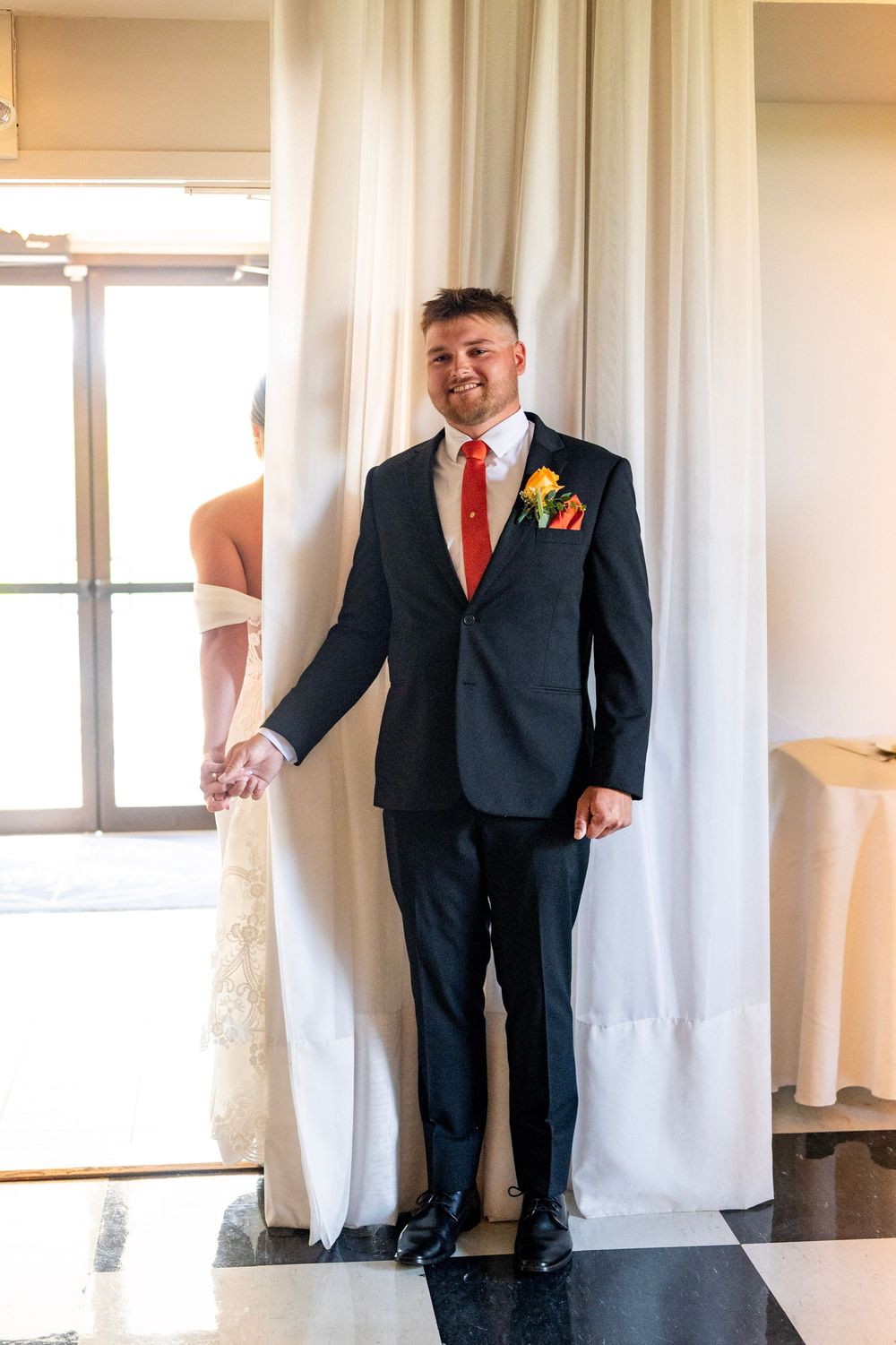 Wedding couple holds hands behind white curtain in formal attire at indoor venue.